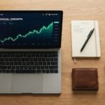 Laptop displaying financial growth chart next to a notebook and coffee on desk.